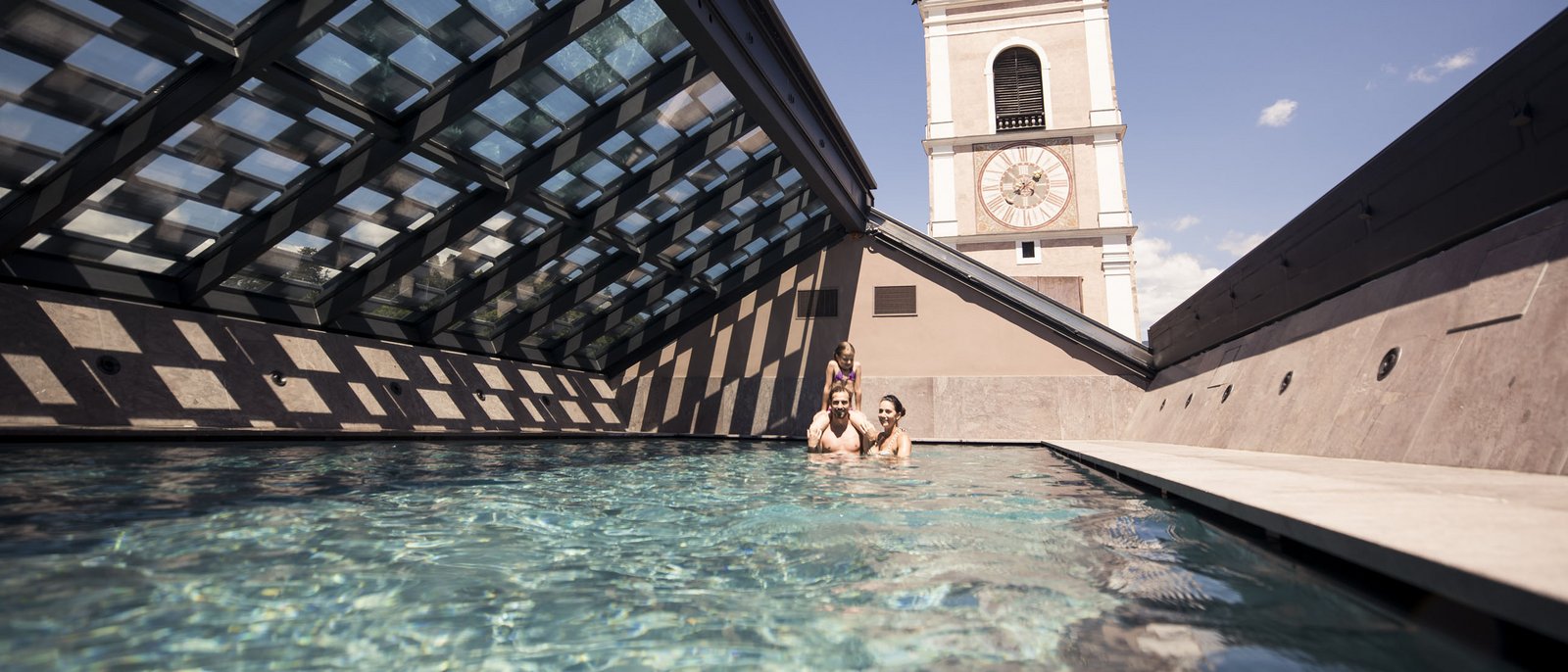 Beim Schwimmen dem Himmel ganz nah sein – im Hotel Lamm! Familie schwimmt in Dachpool mit Blick auf Kirchturm bei klarem Himmel
