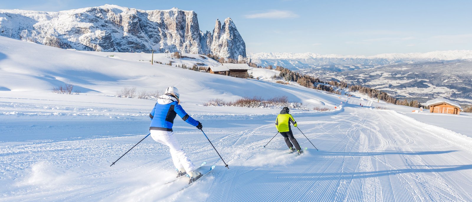 Correte sull’Alpe di Siusi a scoprire lo snowpark più grande dell’Alto Adige! Due sciatori su una pista soleggiata con panorama montano sullo sfondo