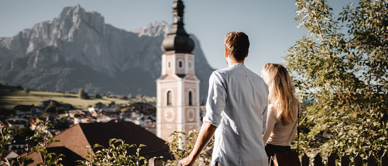 Den Frühling auf der Seiser Alm erleben – jetzt! Paar blickt auf Bergkirche in malerischem Tal mit Bergen im Hintergrund