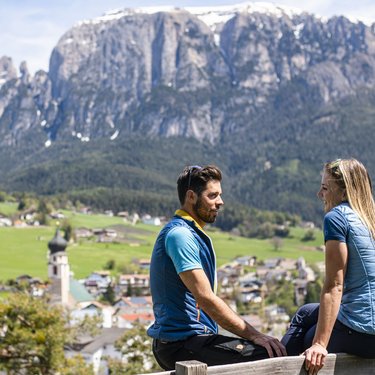 Paar sitzt auf einer Bank mit Blick auf eine Berglandschaft und ein Dorf