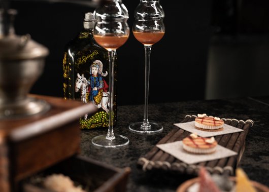 Lampl Stube: il nostro ristorante a Castelrotto Due bicchieri di liquore, una bottiglia con illustrazione colorata e piccoli dolcetti su un vassoio