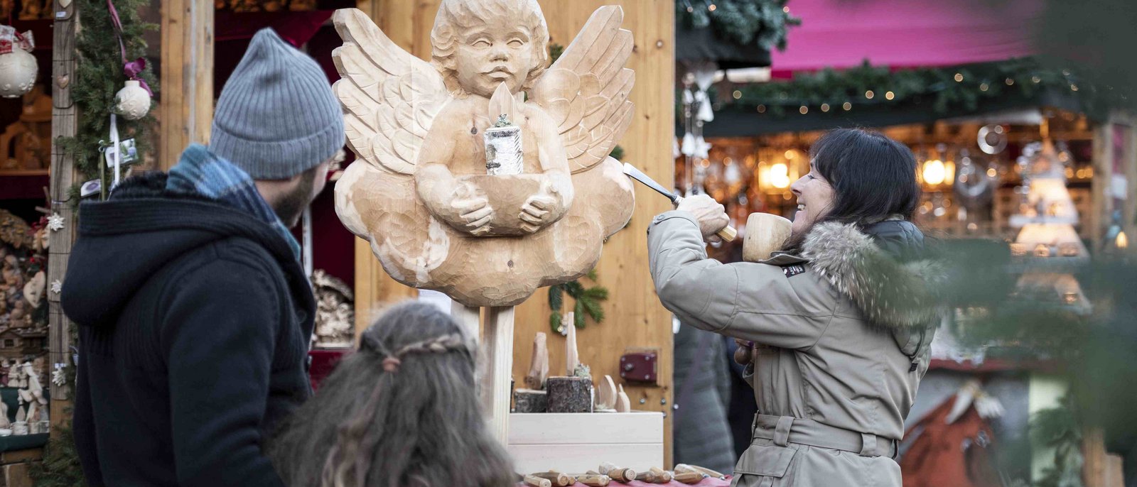 Mercatino di Natale di Castelrotto Donna intaglia una figura d'angelo in legno al mercatino di Natale, famiglia osserva