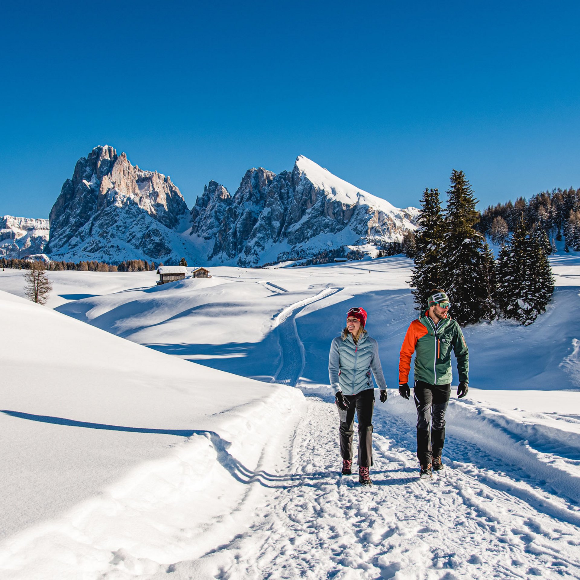 Zwei Wanderer auf verschneitem Weg mit Bergkulisse bei strahlend blauem Himmel