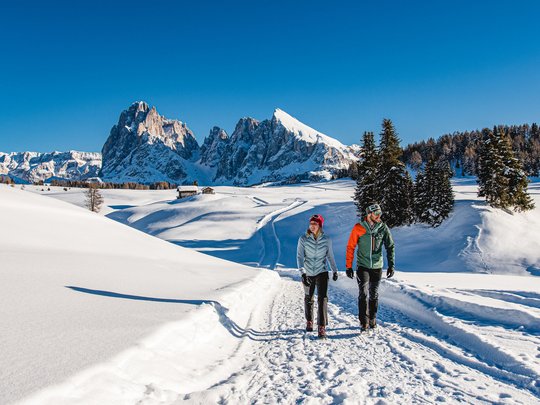 Due escursionisti camminano su sentiero innevato con montagne sullo sfondo e cielo blu